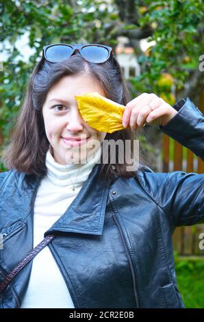 Porträt einer jungen Frau mit kurzen braunen Haaren in schwarz Lederjacke und Sonnenbrille, die ihr Auge mit einem gelben Blatt bedeckt Stockfoto