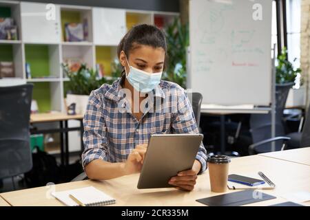 Zurück ins Büro. Junge Frau, weibliche Büroangestellte trägt eine schützende Gesichtsmaske mit einem digitalen Tablet, während sie im Büro sitzt. Geschäft und Arbeit während und covid 19 Ausbruch, soziale Distanzierung Stockfoto