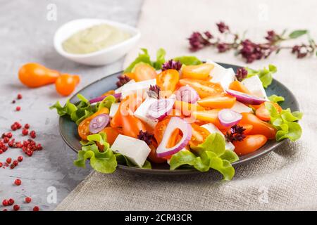Vegetarischer Salat mit frischen Traubentomaten, Feta-Käse, Salat und Zwiebel auf blauer Keramikplatte auf grauem Beton-Hintergrund und Leinentextilien. Seite V Stockfoto