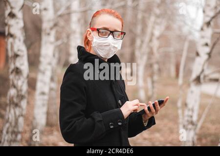 Coronavirus- und Gesundheitskonzept mit Platz für Text. Junge Frau in Maske hält Telefon in der Hand im Park und schaut auf die Kamera. Stockfoto