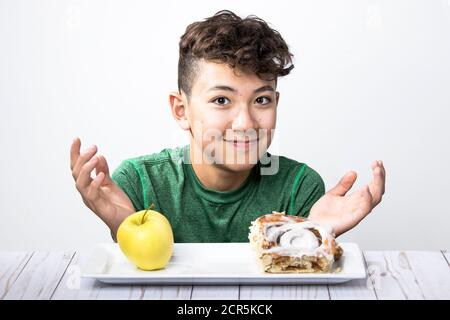 Ein Konzeptfoto eines Teenagers, der versucht, sich zwischen einem gesunden Apfel und einer süßen Zimtrolle zu entscheiden. Stockfoto