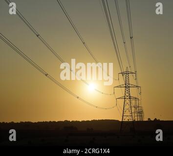 Elektrische Stromleitungen, die bei Sonnenaufgang zu Pylonen/Türmen führen. Stockfoto