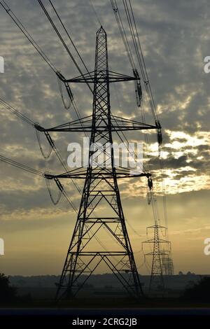 Strom Pylon Übertragung Turm Silhouette bei Sonnenaufgang mit elektrischen Oberleitungen, die zu Pylonen in der Ferne über Landschaft. Stockfoto