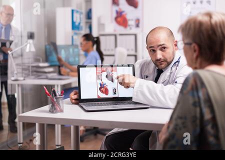 Arzt, der während der Konsultation auf eine ältere Frau blickt und auf ein Herzdiagramm im Krankenhausbüro zeigt. Krankenschwester in blauer Uniform hält Röntgenbild. Stockfoto