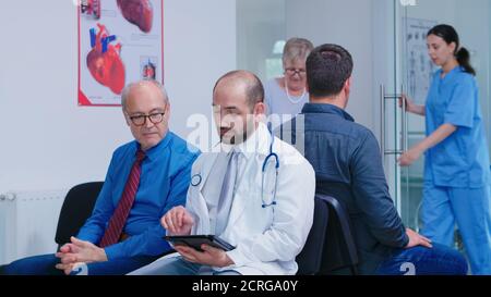 Arzt im weißen Mantel mit Stethoskop Diagnose mit altem Mann im Krankenhaus Wartebereich besprechen. Krankenschwester hilft älteren Frau mit Gehrahmen. Mann wartet auf ärztliche Untersuchung. Stockfoto