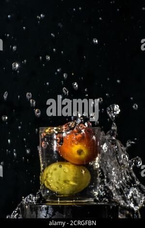 Spritzer rote Tomaten und gelbe Zitrone tauchen in Glas Von Wasser mit schwarzem Hintergrund Stockfoto