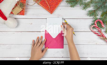 Das junge Mädchen schrieb Brief an den Weihnachtsmann und legt sie in einen Umschlag, flach lag. Weihnachtsdekorationen und Geschenkbox auf Holztisch, Draufsicht. Konz Stockfoto