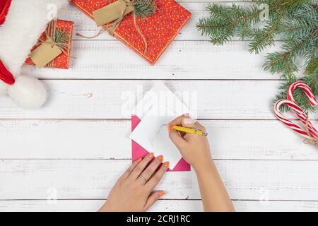 Das junge Mädchen schrieb Brief an den Weihnachtsmann und legt sie in einen Umschlag, flach lag. Weihnachtsdekorationen und Geschenkbox auf Holztisch, Draufsicht. Konz Stockfoto