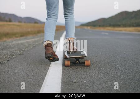 Crop Foto von Frauen Beine in Jeans auf Longboard auf Straße mit Bergen Hintergrund Stockfoto