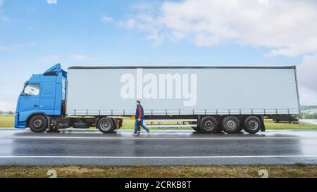 LKW-Fahrer überquert die Straße im ländlichen Raum und steigt in seine Blue Long Haul Semi-Truck mit Cargo Trailer befestigt. Logistikunternehmen Umzug Stockfoto