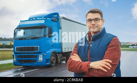 Happy Professional Young Truck Driver kreuzt Arme und Smiles an der Kamera. Hinter ihm geparkt Blue Long Haul Semi-Truck mit Cargo Trailer Stockfoto