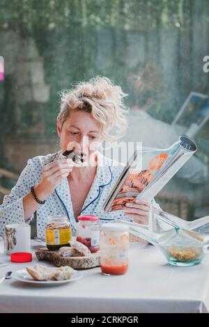 Blonde junge reife Frau in Pyjamas zu Hause in der Frühstückszeit, Lesen einer Zeitschrift und Essen einen Toast. Stockfoto