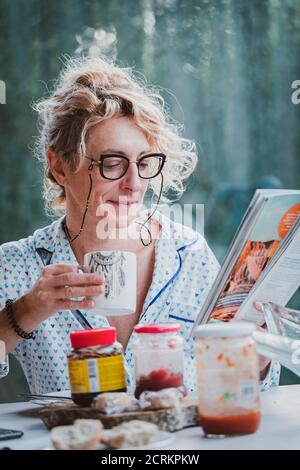 Blonde junge reife Frau mit Brille im Pyjama zu Hause in der Frühstückszeit, Lesen einer Zeitschrift und mit einer Tasse Kaffee Stockfoto