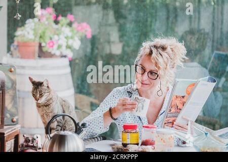 Blonde junge reife Frau mit Brille im Pyjama zu Hause in der Frühstückszeit, Lesen einer Zeitschrift und mit einer Tasse Kaffee und Katze. Stockfoto