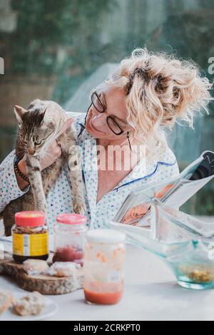 Blonde junge reife Frau mit Brille im Pyjama zu Hause in der Frühstückszeit, Lesen einer Zeitschrift und einen Moment mit seiner Katze. Stockfoto