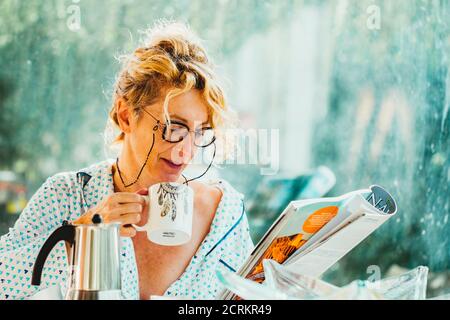 Blonde junge reife Frau mit Brille im Pyjama zu Hause in der Frühstückszeit, Lesen einer Zeitschrift und mit einer Tasse Kaffee Stockfoto