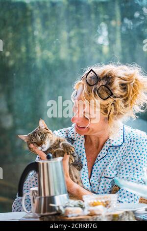 Blonde junge reife Frau mit Brille im Pyjama zu Hause in der Frühstückszeit, Lesen einer Zeitschrift und einen Moment mit seiner Katze. Stockfoto