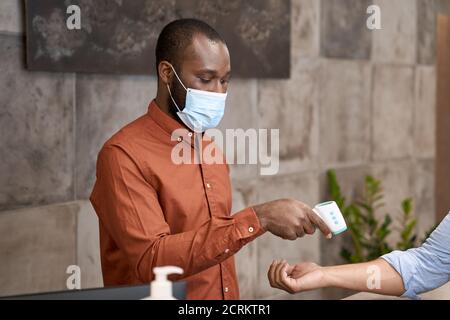 Afrikanischer männlicher Rezeptionist trägt medizinische Maske mit Infrarot-Thermometer für Körpertemperatur des Büroangestellten messen Stockfoto
