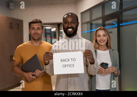 Covid 19 und Business, junge fröhliche afrikanische Büroangestellte zeigt Papier mit Text ZURÜCK INS BÜRO an der Kamera und lächelt, während er mit ihm steht Stockfoto