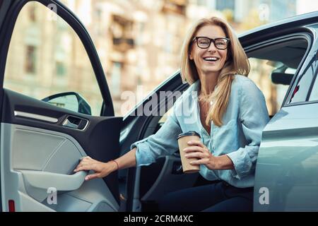Glückliche attraktive Frau oder Geschäftsfrau trägt Brillen halten Tasse Von Kaffee und aus ihrem modernen Auto aussteigen Stockfoto