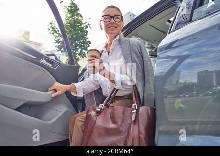 Schöne lächelnde Frau mittleren Alters mit Handtasche halten Tasse Von Take Away Kaffee beim Aussteigen aus ihrem modernen Auto Stockfoto