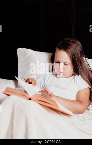 Das Kind sitzt auf dem Bett, bedeckt mit einer leichten Decke, und liest das Buch, das er auf seinem Schoß hält, und dreht mit der Hand die Seite um Stockfoto