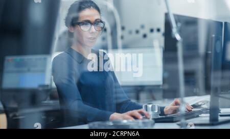 Durch Glas Geschossen. Ingenieurin, die am Personal Computer arbeitet. Sie arbeitet im Büro in der modernen Fabrik mit High-Tech-Maschinen. Stockfoto