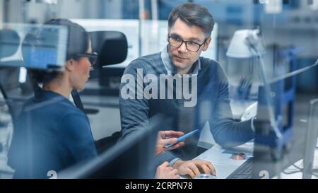 Shot Through Glass in Modern Factory: Weibliche Projektleiterin spricht mit einem männlichen Industrieingenieur, der am Personal Computer arbeitet. Moderne High-Tech-Technik Stockfoto