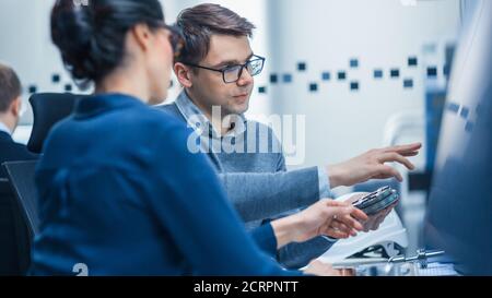 Moderne Fabrik: Männlicher Industrieingenieur erklärt weiblichen Projektleiters Funktionen des Maschinenteil, das es mit einem auf Computerbildschirm vergleicht Stockfoto