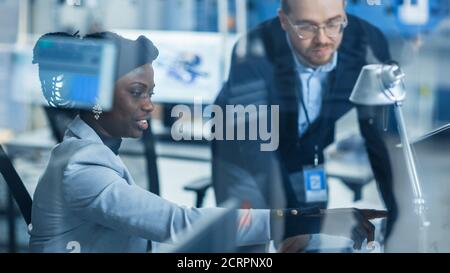 Shot Through Glass in Modern Electronics Factory: Weibliche Vorgesetzte spricht mit einem männlichen Elektroingenieur, der am Computer arbeitet. PCB-Design Stockfoto