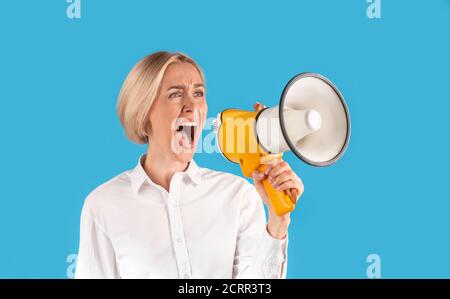 Aufgeregte blonde Frau in formeller Kleidung schreiend in Megaphon auf blauem Studiohintergrund, Panorama Stockfoto