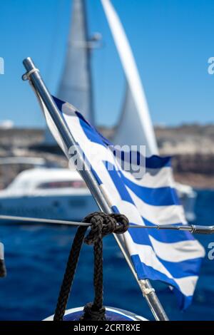 Griechische Flagge flattert im Wind auf einer Segelyacht, die auf dem blauen Meer gegen den blauen Himmel segelt. Verschwommenes Segelboot im Hintergrund Stockfoto
