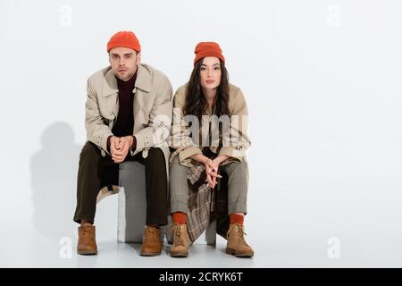 Junge Brünette Frau im Trenchcoat sitzen mit geballten Händen In der Nähe trendy Mann in Mütze Hut auf weiß Stockfoto