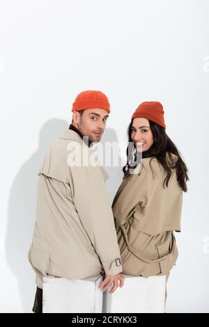 Trendige Paar in Beanie Hüte und Trenchcoats sitzen und Blick auf die Kamera auf weiß Stockfoto