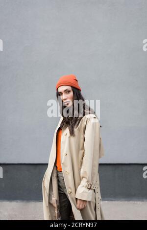 Junge trendige Frau in Trenchcoat und Beanie Hut suchen Bei der Kamera Stockfoto