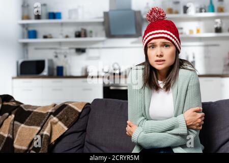 Eiskalte Frau in gestricktem Hut umarmt sich, während sie sitzt Kalte Küche Stockfoto