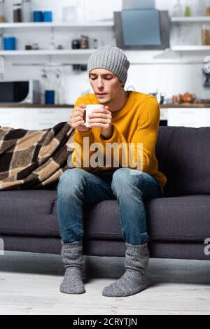 Junger Mann in Strickpullover, Hut und Socken, der auf heißen Tee bläst, während er auf dem Sofa in der kalten Küche sitzt Stockfoto