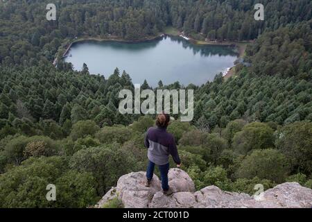 Männlicher Wanderer, der am Rand der Klippe steht und einen Wald und einen See bewundert Stockfoto