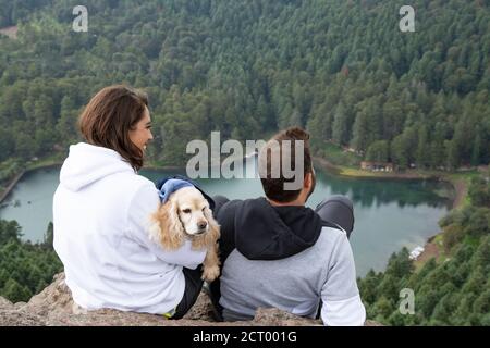 Ein junges mexikanisches Paar und ein Hund, der den Blick auf einen See und einen Wald bewundert Stockfoto