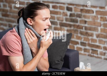 Kranke junge Mann in warmen Schal mit Kehlspray während Berühren des Halses Stockfoto