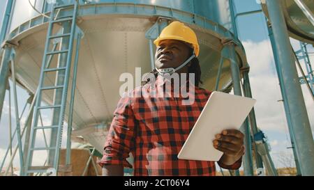 Junger schöner afrikanischer Mann, Ingenieur mit Schutzhelm mit Tablet vor dem Silosystem. Hochwertige Fotos Stockfoto