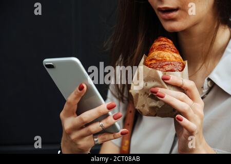 Zugeschnittenes Bild einer Frau auf der Straße mit Croissant, Blick auf ihr Telefon. Stockfoto