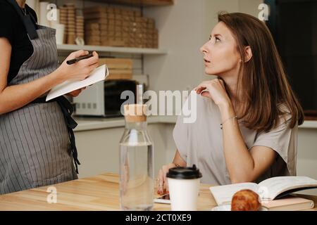 Kellnerin nimmt Auftrag von reifer Frau, sitzen hinter dem Tisch. Stockfoto