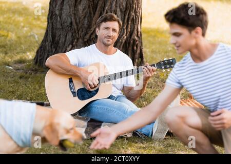 Selektiver Fokus des Mannes unter Baum sitzend und akustisch spielend Gitarre beim Blick auf Teenager Sohn und Golden Retriever Stockfoto