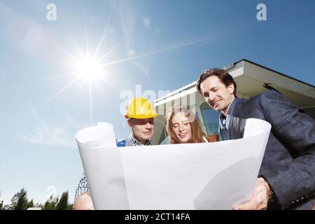 Arbeiter zeigt Bauplan für junge Paar außerhalb Haus unter Bausonne scheint Stockfoto