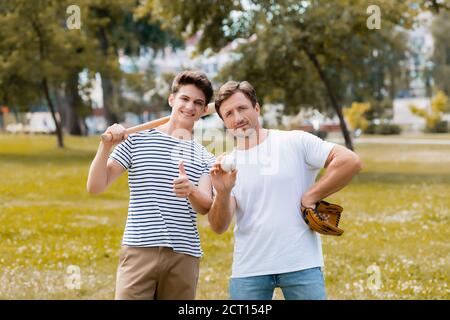 Teenager Sohn mit Softball Fledermaus zeigt Daumen in der Nähe Vater In Leder Handschuh halten Ball Stockfoto