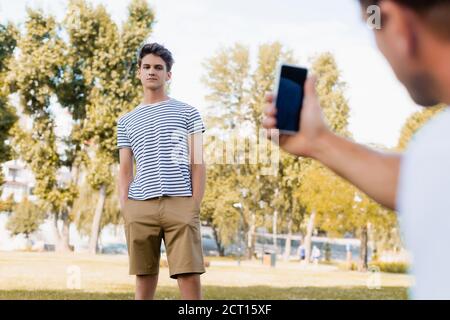 Selektiver Fokus des Teenagers Jungen stehend mit Händen in den Taschen Während Vater Foto auf dem Smartphone im Park Stockfoto