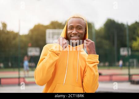 afroamerikanisch lächelnder schwarzer Mann mit Kapuze auf dem Kopf, gelb gekleidet hoodie außerhalb glücklich emotionale afro Modell Kerl zu Fuß Skate Park Sommertag Fre Stockfoto
