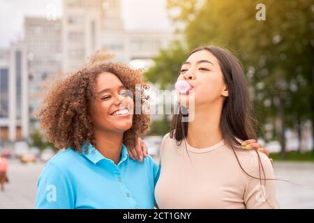 afroamerikanische Frau lachend, während asiatische Freund aufblasende Kaugummi. Nahaufnahme von multiethnischen Freunden, die die Straße im Freien genießen. Mädchen lachend an Stockfoto