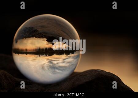 Blick durch einen Lensball mit schönen Reflexen des orangen Sonnenuntergangs, Himmel und Wasser an einem Sommerabend. Stockfoto